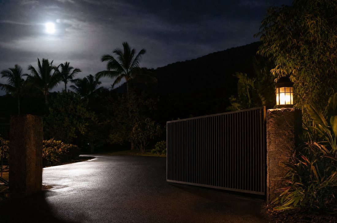A dark metal sliding driveway gate standing half open at night, a warm lamp on the stone post beside it lighting the wet asphalt.